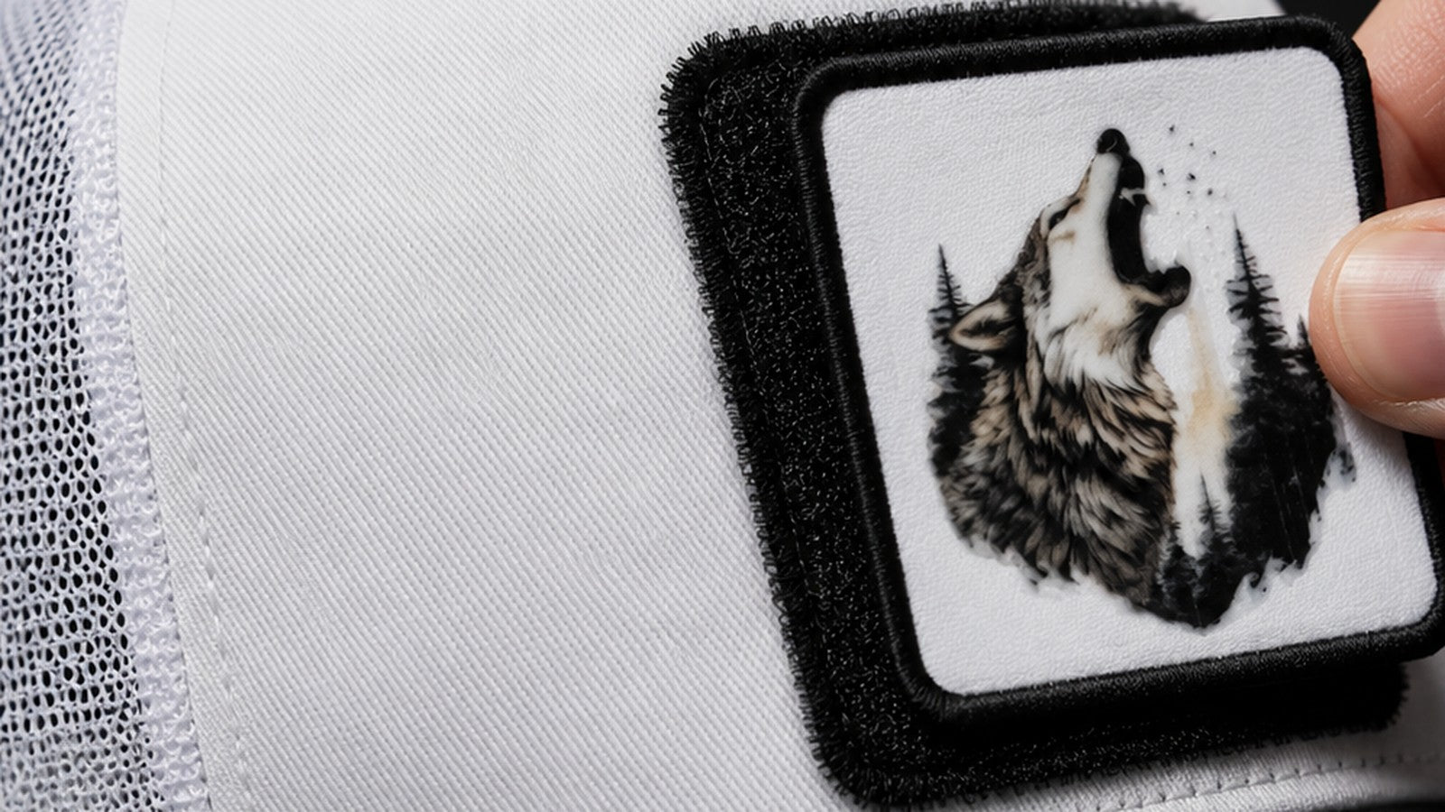 Close-up of a hand lifting a wolf patch off the hook-and-loop panel on the front of a white BlackBork trucker cap